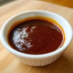 Close-up of thick, dark red homemade bbq sauce glistening in a small white bowl on a wooden surface.
