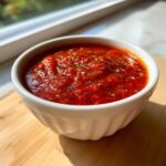 Close-up of rich, vibrant red pizza sauce topped with dried herbs in a small white bowl.