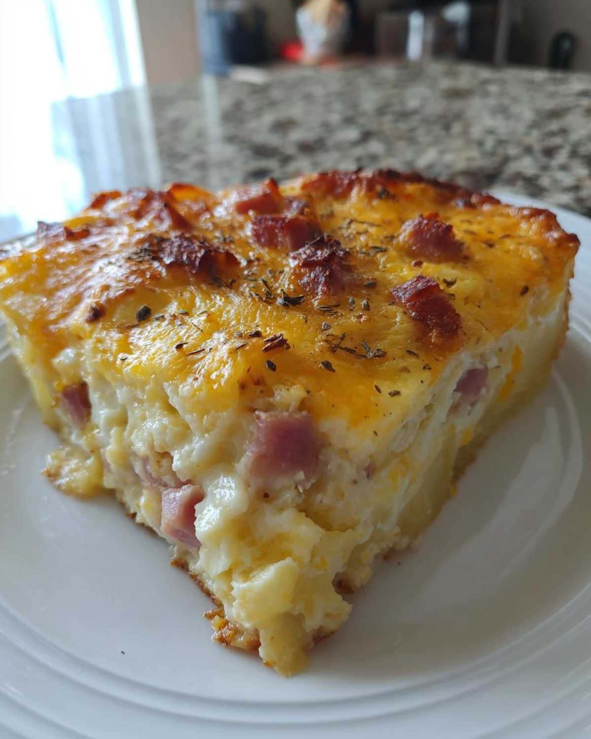A square slice of baked scalloped potatoes and ham casserole topped with melted cheese and herbs.