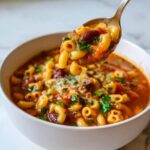 A spoonful of rich pasta fagioli soup, showing elbow macaroni, kidney beans, and fresh parsley, lifted from a white bowl.