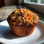 A single, perfectly baked morning glory muffin topped with nuts and seeds, sitting on a white plate.