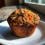 A single, perfectly baked morning glory muffin topped with nuts and seeds, sitting on a white plate.