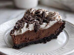 A close-up of a decadent slice of chocolate mousse pie with a dark crust, chocolate filling, and whipped cream topping.