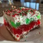 Close-up of a festive slice of christmas poke cake showing red and green gelatin layers, whipped topping, and sprinkles.