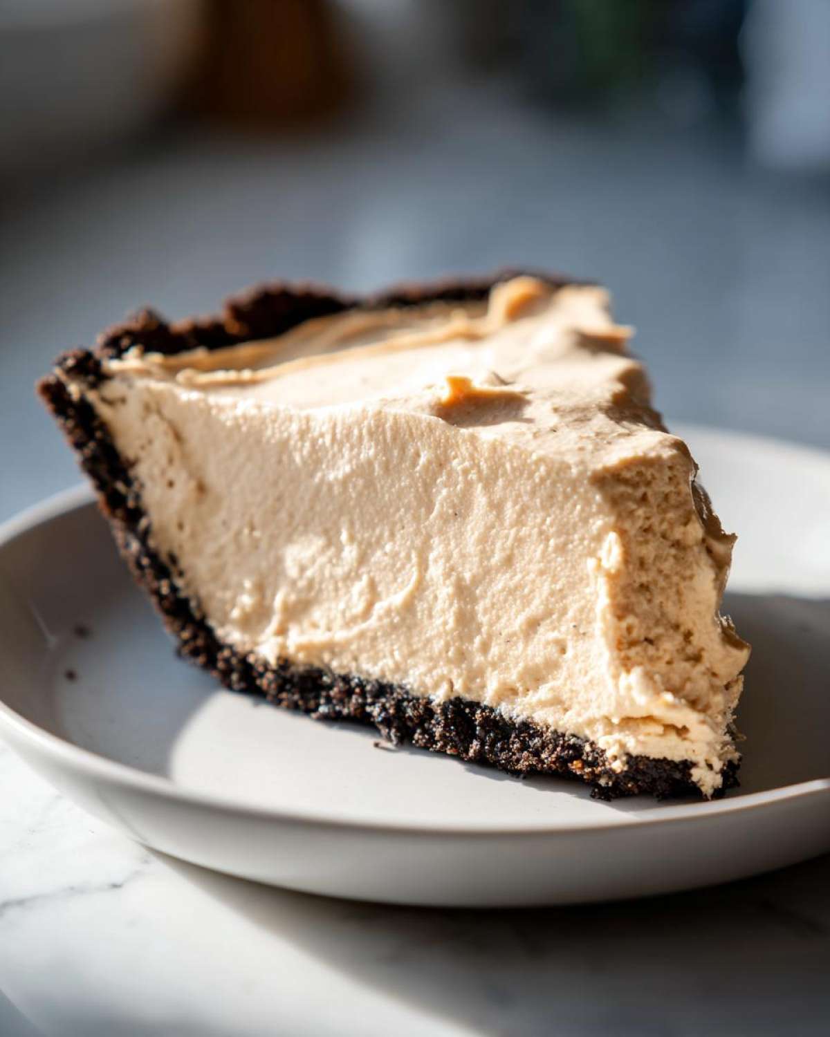 A thick slice of no bake peanut butter pie featuring a creamy filling and dark chocolate cookie crust on a light plate.
