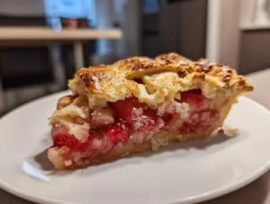 A close-up of a perfect slice of strawberry rhubarb pie featuring a golden lattice crust and juicy red filling.