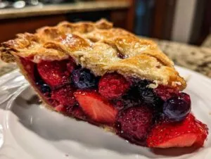 Close-up of a thick slice of triple berry pie showing strawberries, blueberries, and raspberries under a flaky, golden lattice crust.