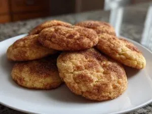 A close-up of several soft, golden snickerdoodle cookies piled on a white plate, coated in cinnamon sugar.