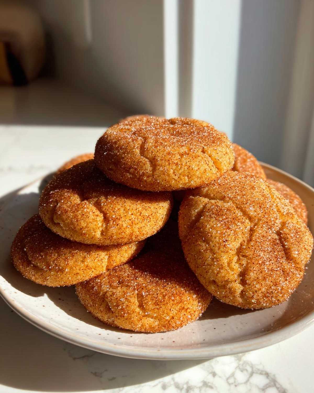 A close-up stack of soft, golden snickerdoodle cookies generously coated in cinnamon sugar, sitting on a speckled plate.
