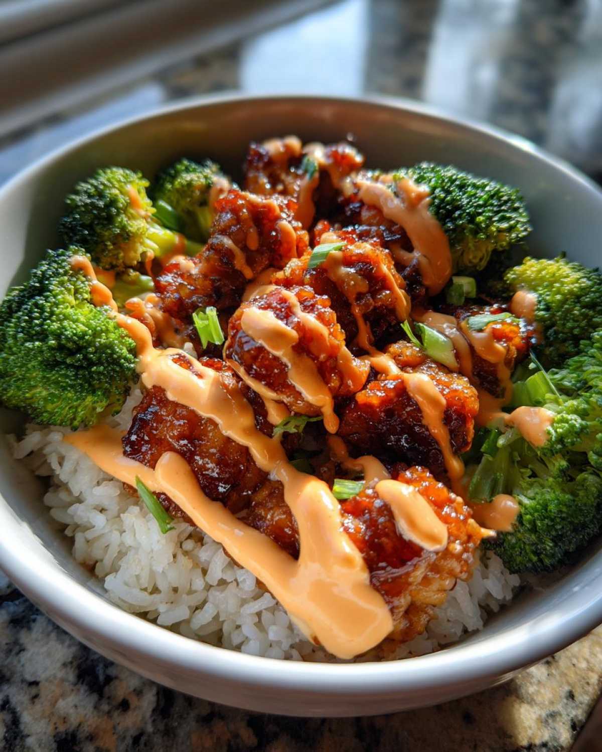 Close-up of a sticky chicken rice bowl featuring glazed chicken pieces, steamed broccoli, and a drizzle of orange sauce.