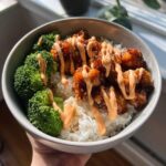 A close-up of a sticky chicken rice bowl topped with bright green broccoli florets and drizzled with a creamy orange sauce.