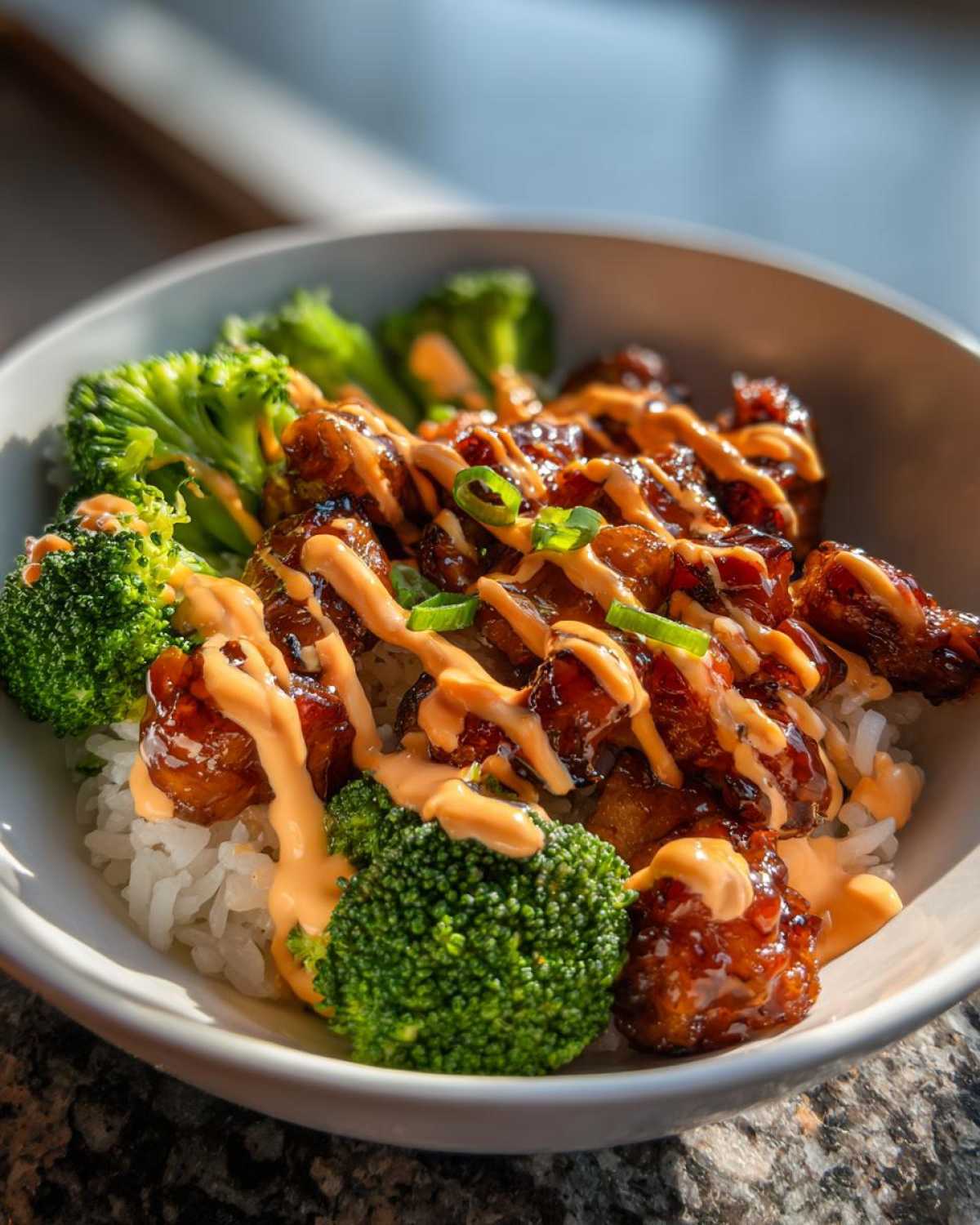 Close-up of a sticky chicken rice bowl featuring glazed chicken pieces, bright green broccoli florets, and a drizzle of spicy orange sauce.