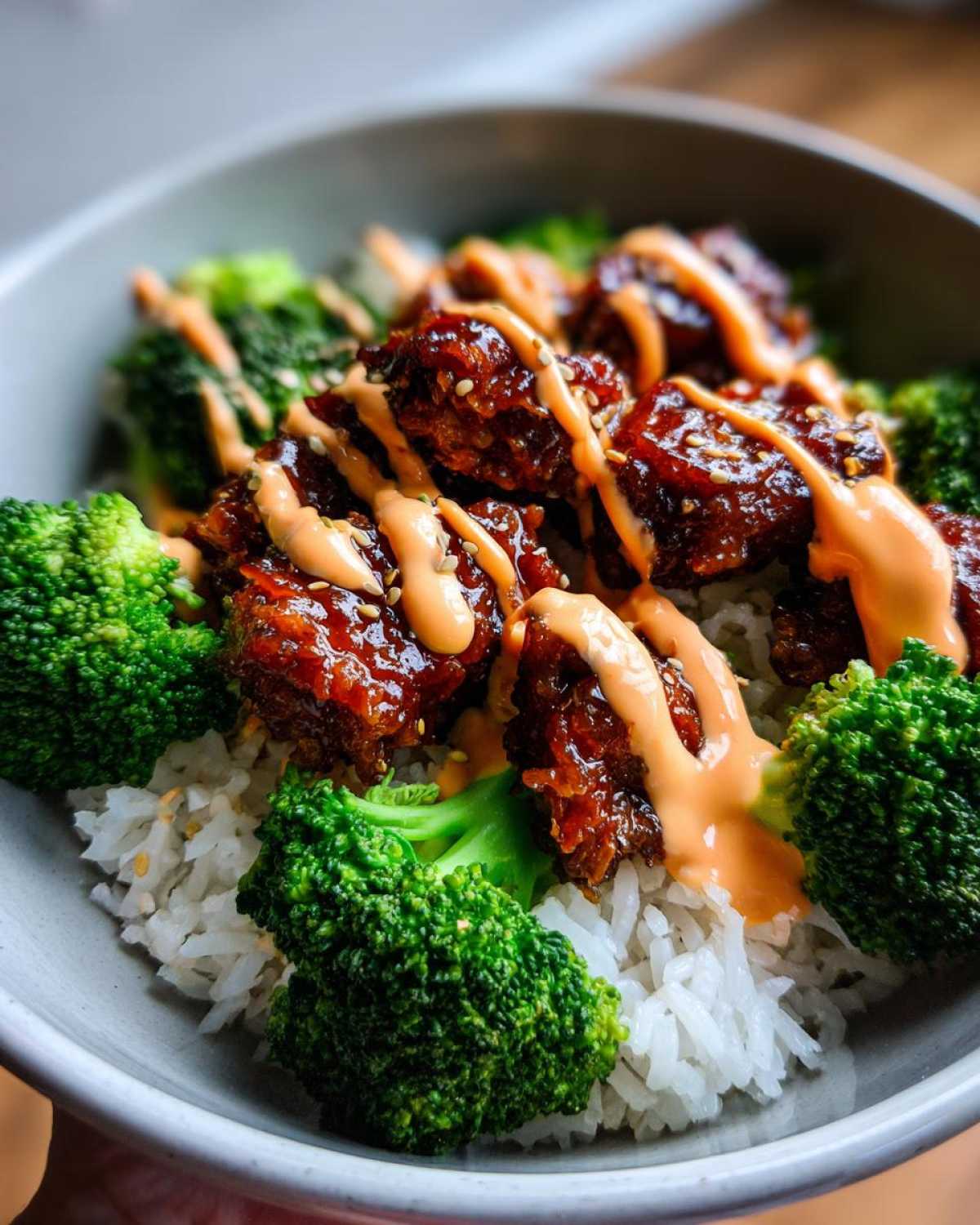 Close-up of a sticky chicken rice bowl featuring glazed chicken pieces, bright green broccoli florets, and a drizzle of orange sauce.