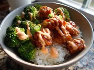 Close-up of a sticky chicken rice bowl featuring glazed chicken pieces, bright green broccoli, and a drizzle of spicy mayo sauce.