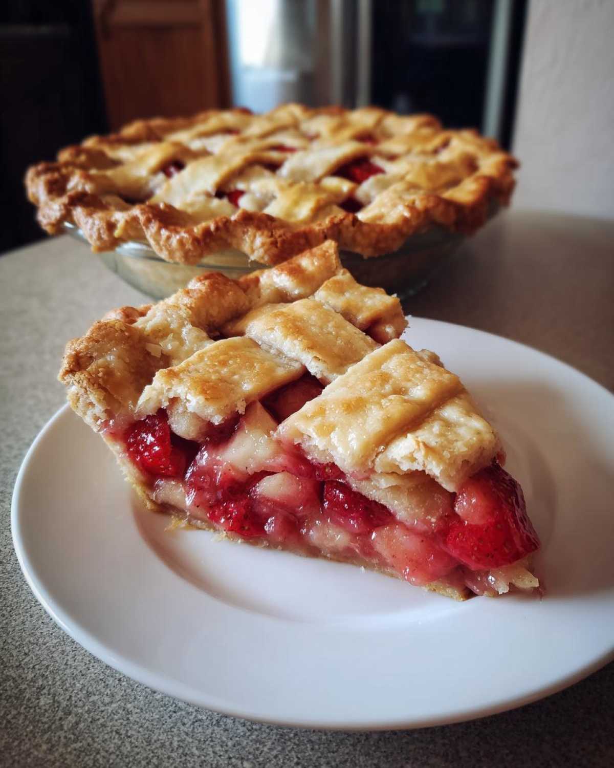 A close-up of a slice of strawberry rhubarb pie with a golden lattice crust on a white plate.
