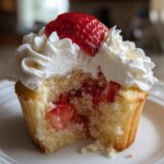Close-up of a strawberry shortcake cupcake with a bite taken out, showing strawberry filling and whipped cream topping.