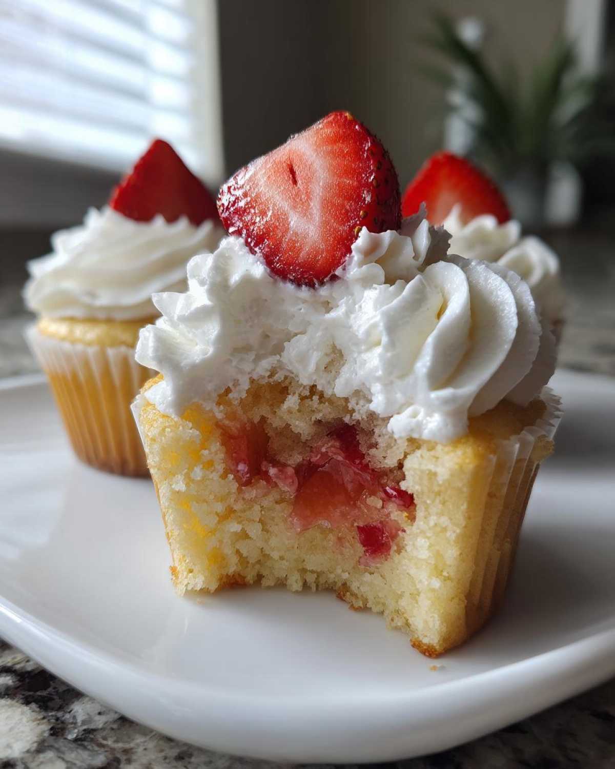 Close-up of a strawberry shortcake cupcake with a bite taken out, revealing strawberry filling and topped with whipped cream.