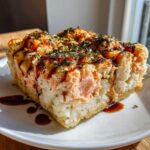 A square slice of baked salmon sushi bake casserole topped with sauce and seaweed flakes.