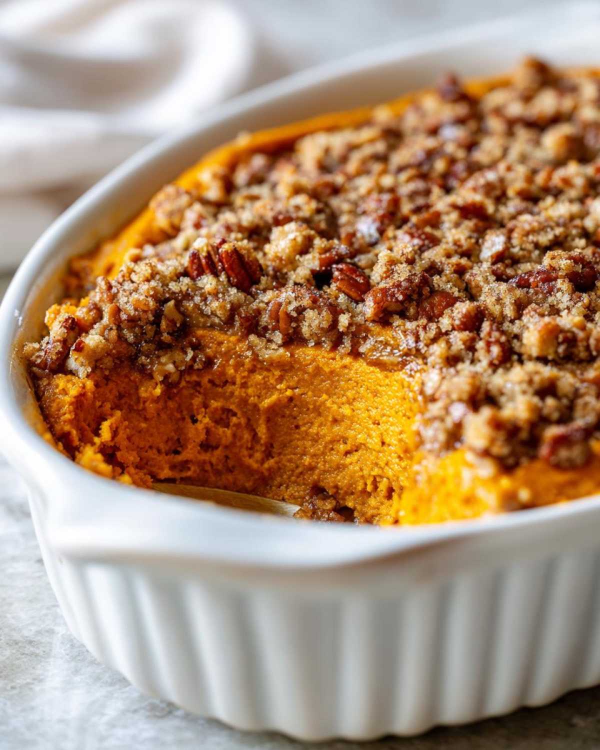 A close-up of a rich, orange sweet potato souffle in a white baking dish, topped with a crunchy pecan streusel.