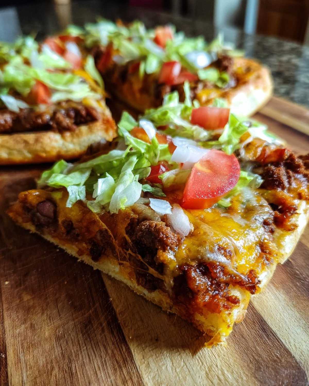 A close-up slice of taco pizza showing seasoned meat, melted cheese, lettuce, tomato, and onion on a wooden board.