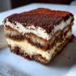 Close-up of a layered slice of tiramisu recipe, showing coffee-soaked ladyfingers, mascarpone cream, and cocoa powder topping.