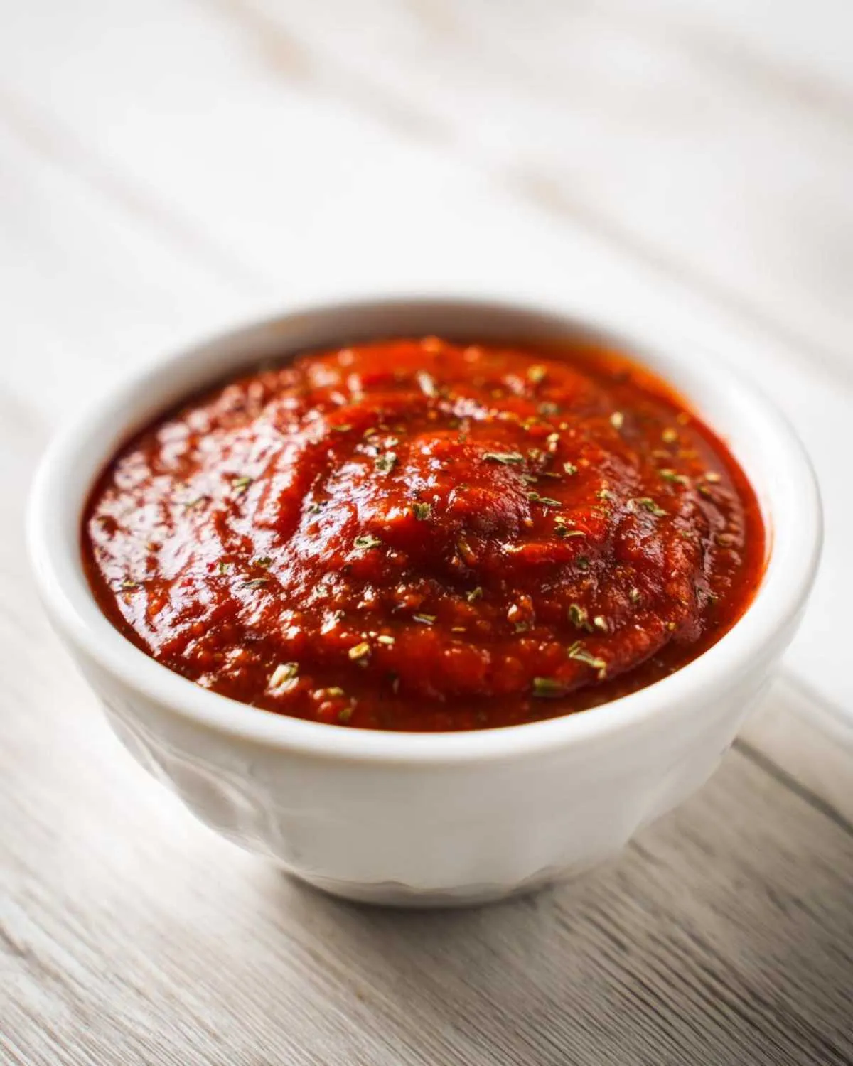 Close-up of thick, vibrant red pizza sauce seasoned with herbs in a small white bowl.