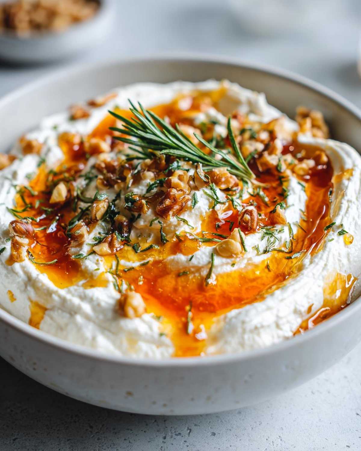 Close-up of a bowl of whipped ricotta dip with hot honey drizzle, chopped walnuts, and a rosemary sprig.