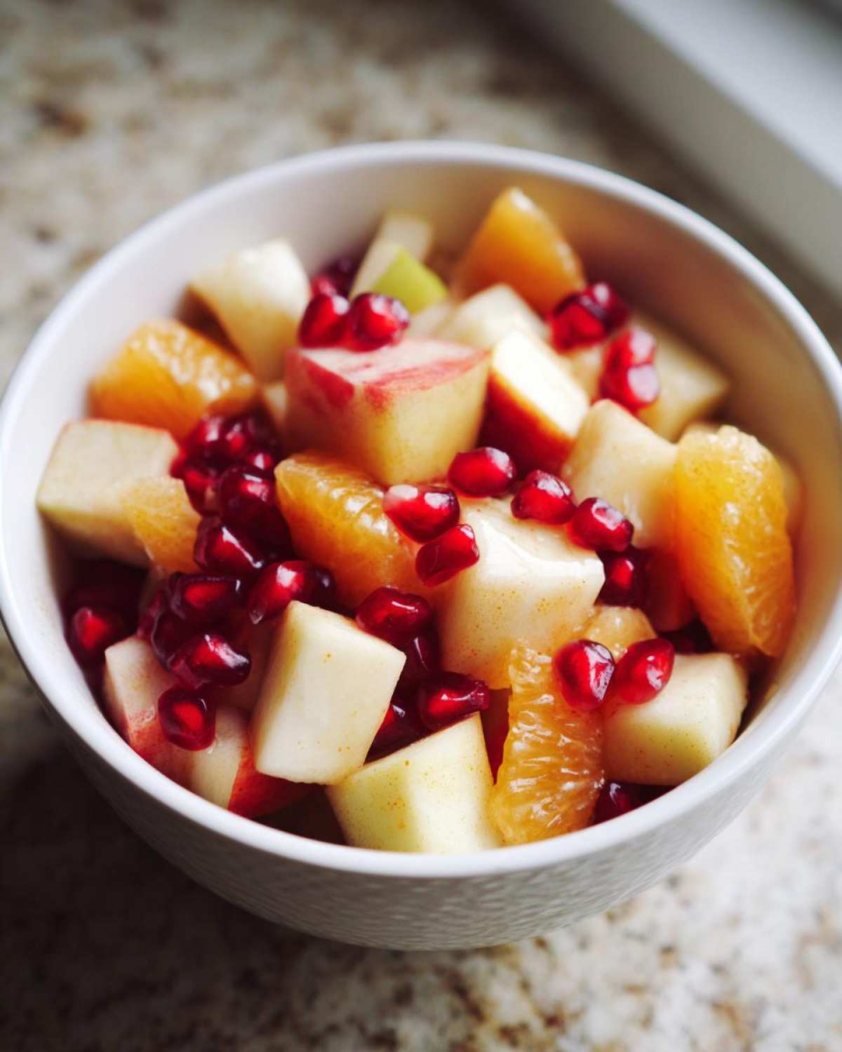 Close-up of a white bowl filled with colorful winter fruit salad featuring diced apples, orange segments, and bright pomegranate seeds.