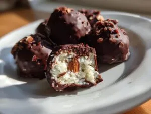 A plate of dark chocolate-covered almond joy truffles, one cut open revealing coconut filling and a whole almond.
