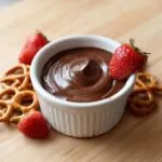 A small white bowl filled with smooth, rich chocolate dip, garnished with fresh strawberries and surrounded by pretzels.
