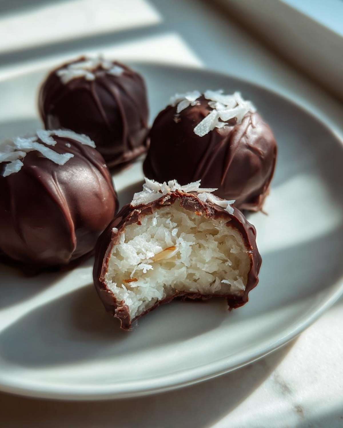 Four chocolate-covered almond joy truffles on a plate, one is bitten showing the coconut filling and almond center.