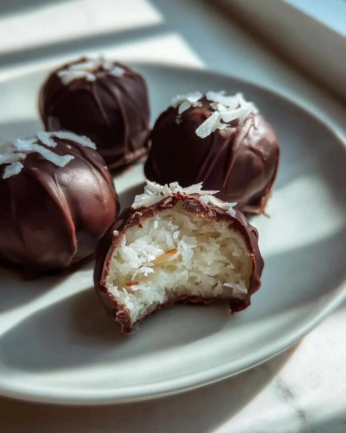 Four chocolate-covered almond joy truffles on a plate, one is bitten showing the coconut filling and almond center.