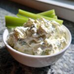 A close-up of a white bowl filled with chunky blue cheese dip, served with celery sticks in the background.