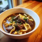 A close-up of a white bowl filled with rich beef noodle soup, featuring tender chunks of beef and wide egg noodles, garnished with parsley.