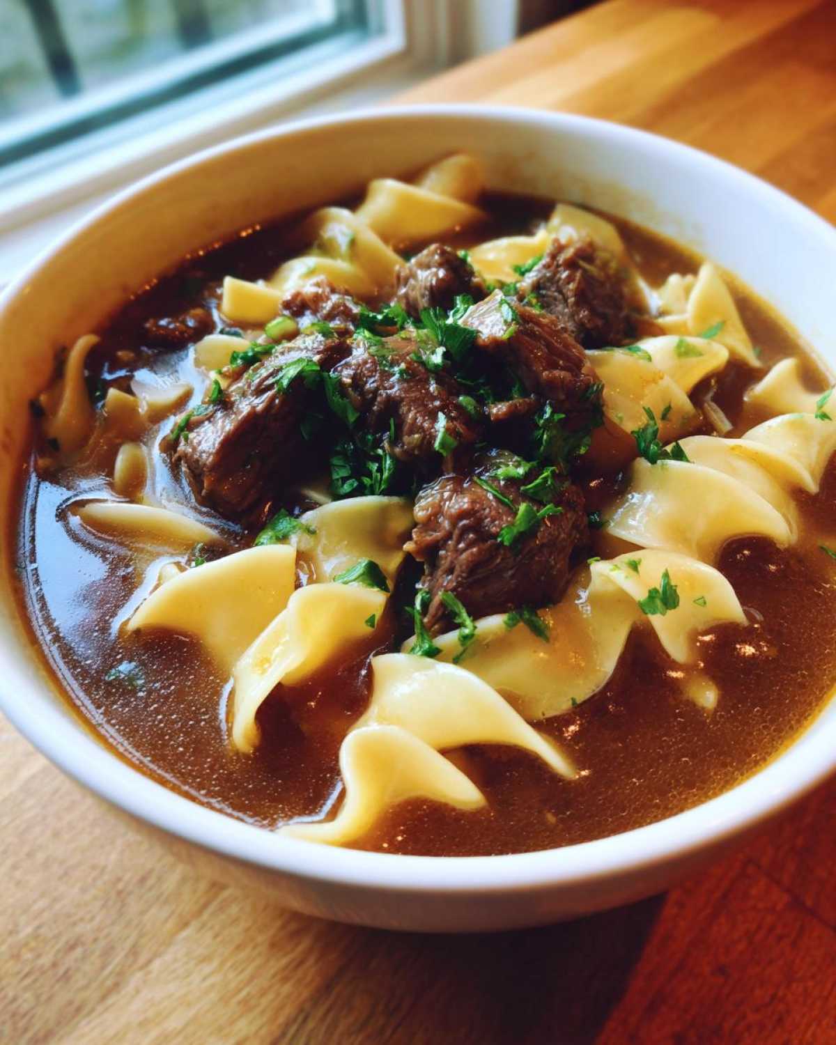 Close-up of a white bowl filled with rich broth, wide egg noodles, and tender chunks of beef in this amazing beef noodle soup recipe.