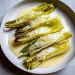 Five pieces of tender braised leeks covered in a light yellow cream sauce on a white plate.