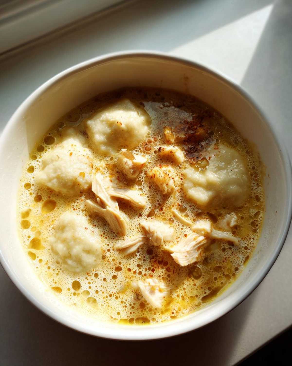 Overhead view of a white bowl filled with rich, yellow broth, fluffy dumplings, and shredded chicken, representing amazing chicken and dumplings.