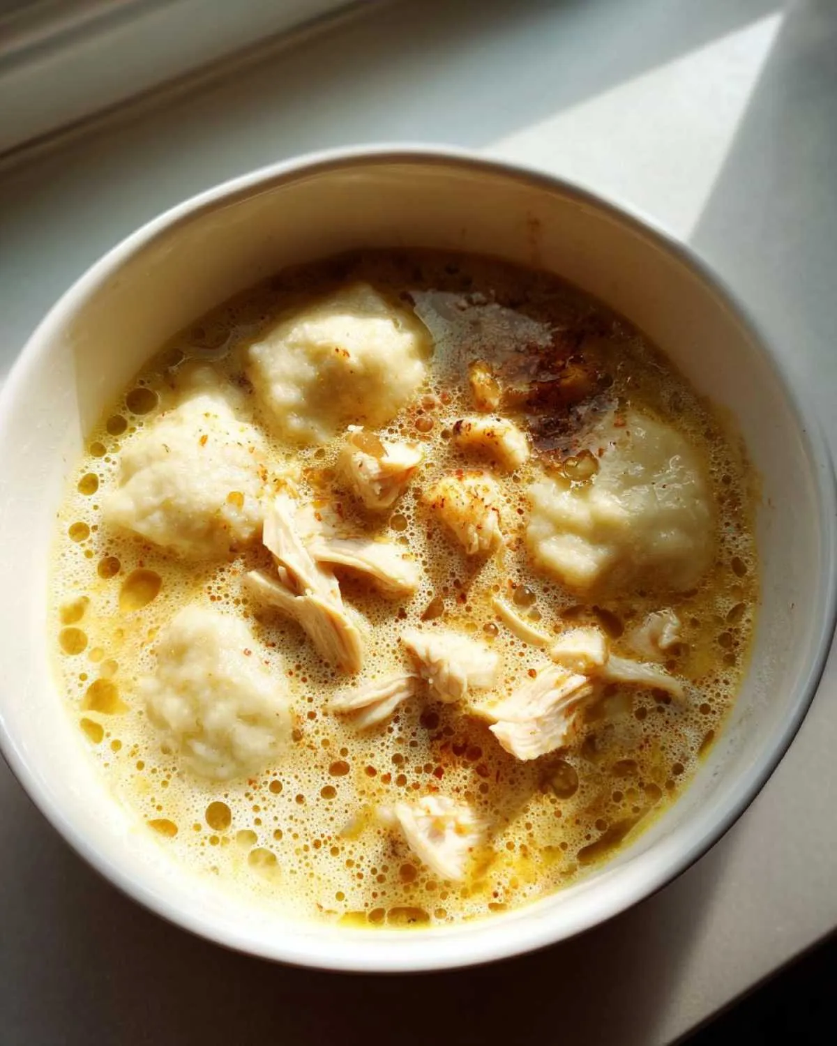 Overhead view of a white bowl filled with rich, yellow broth, fluffy dumplings, and shredded chicken, representing amazing chicken and dumplings.