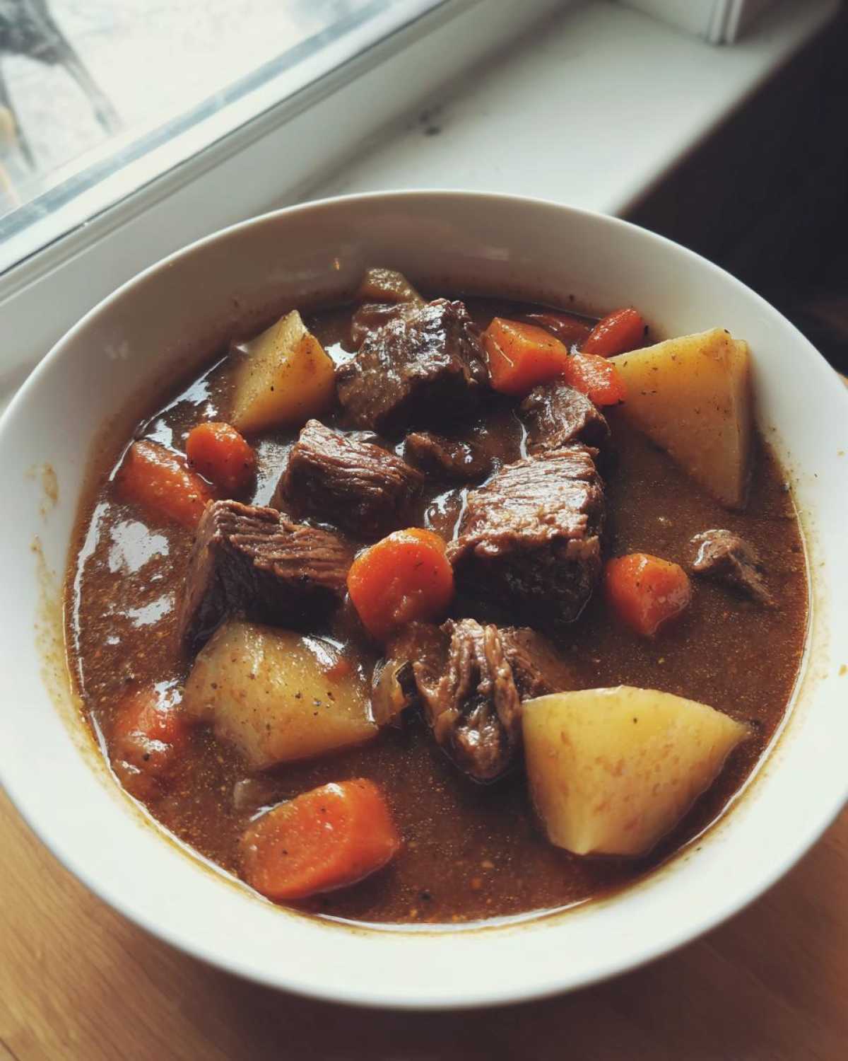 A close-up of a white bowl filled with rich, dark pork stew featuring chunks of meat, potatoes, and carrots.