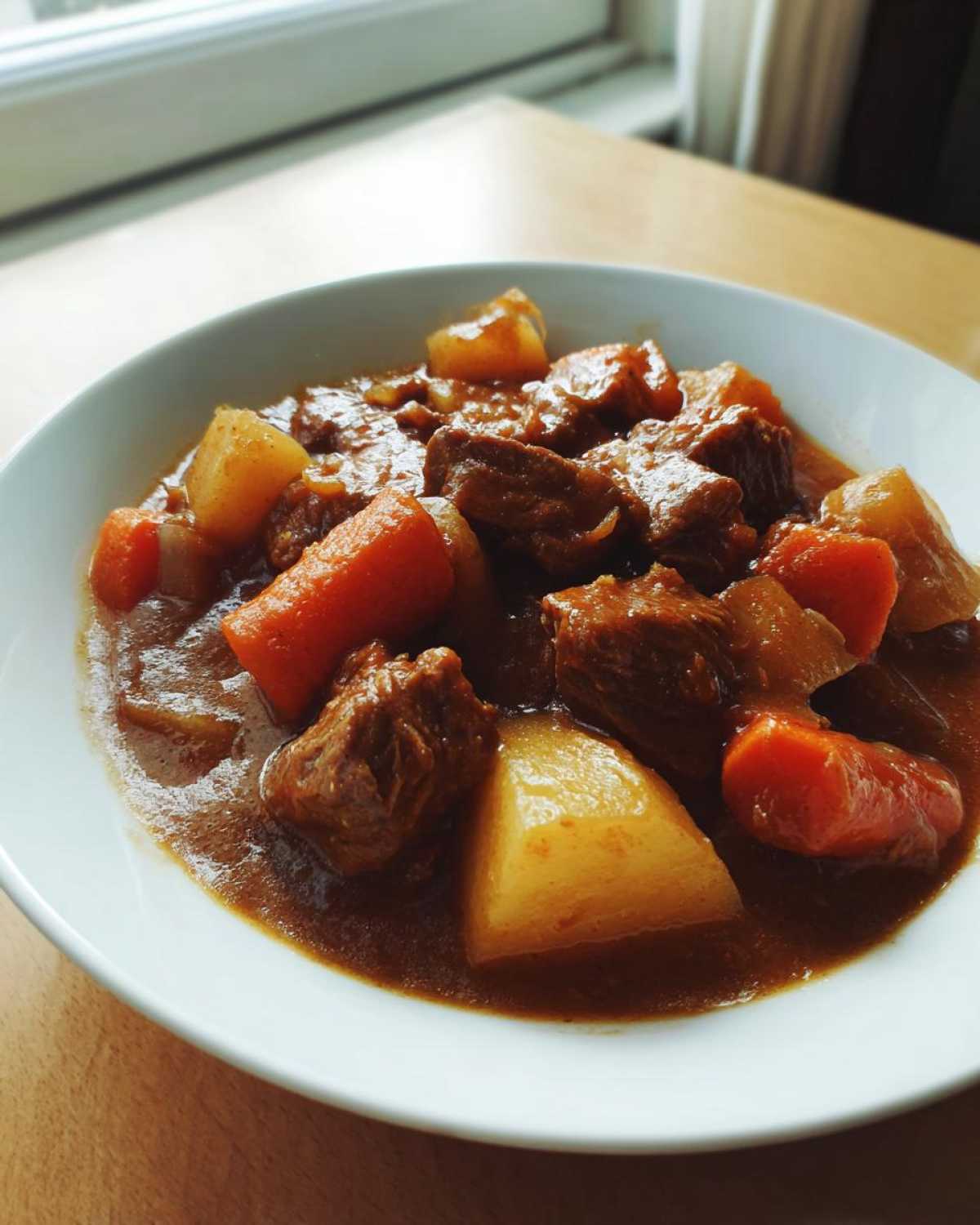 Close-up of a white bowl filled with rich, brown Amazing 3-Hour Pork Stew featuring chunks of meat, carrots, and potatoes.