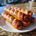 A stack of freshly baked pretzel dogs with coarse salt on a white plate, showing the browned pretzel dough wrapped around the hot dogs.