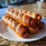 A stack of freshly baked pretzel dogs with coarse salt on a white plate, showing the browned pretzel dough wrapped around the hot dogs.