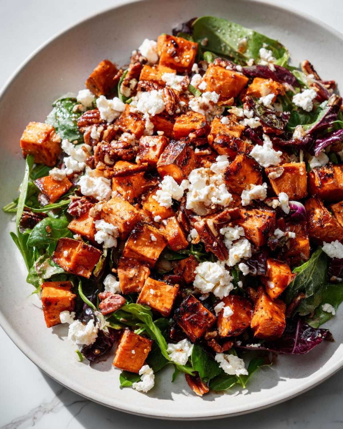 Close-up of a bowl featuring amazing sweet potato salad with roasted sweet potato cubes, crumbled feta cheese, and pecans over mixed greens.