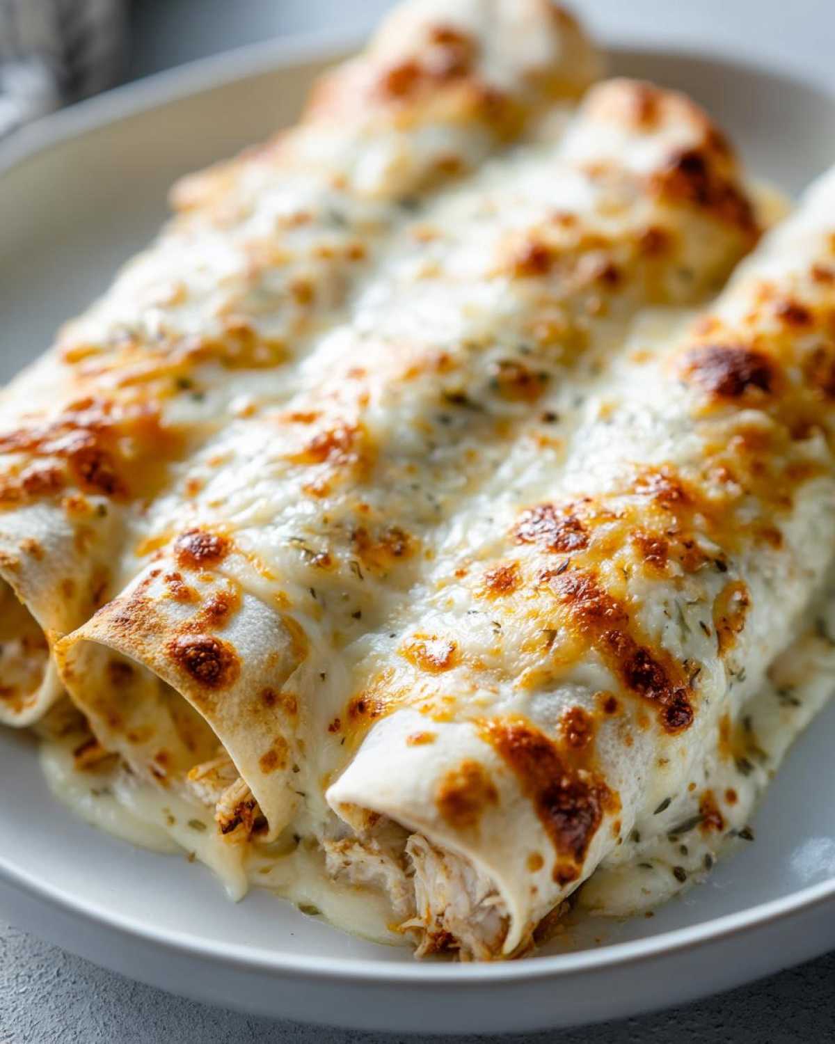 Close-up of three baked white chicken enchiladas covered in melted, bubbly white cheese and herbs.