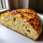 Close-up of a slice of artisan bread showing an open crumb structure and a golden, cheesy, herbed crust.