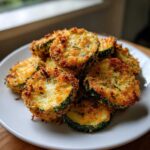 A pile of golden brown, crispy baked parmesan zucchini slices served on a white plate.