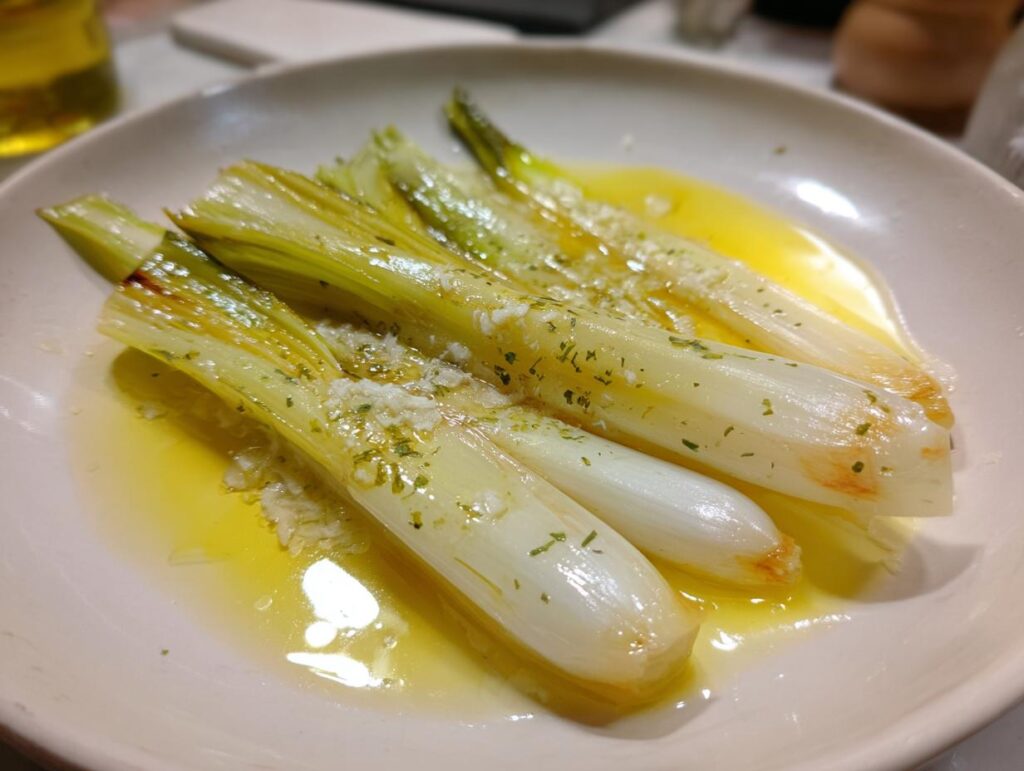 Four tender braised leeks served on a white plate swimming in melted butter, topped with garlic and herbs.