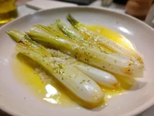 Four tender braised leeks served on a white plate swimming in melted butter, topped with garlic and herbs.