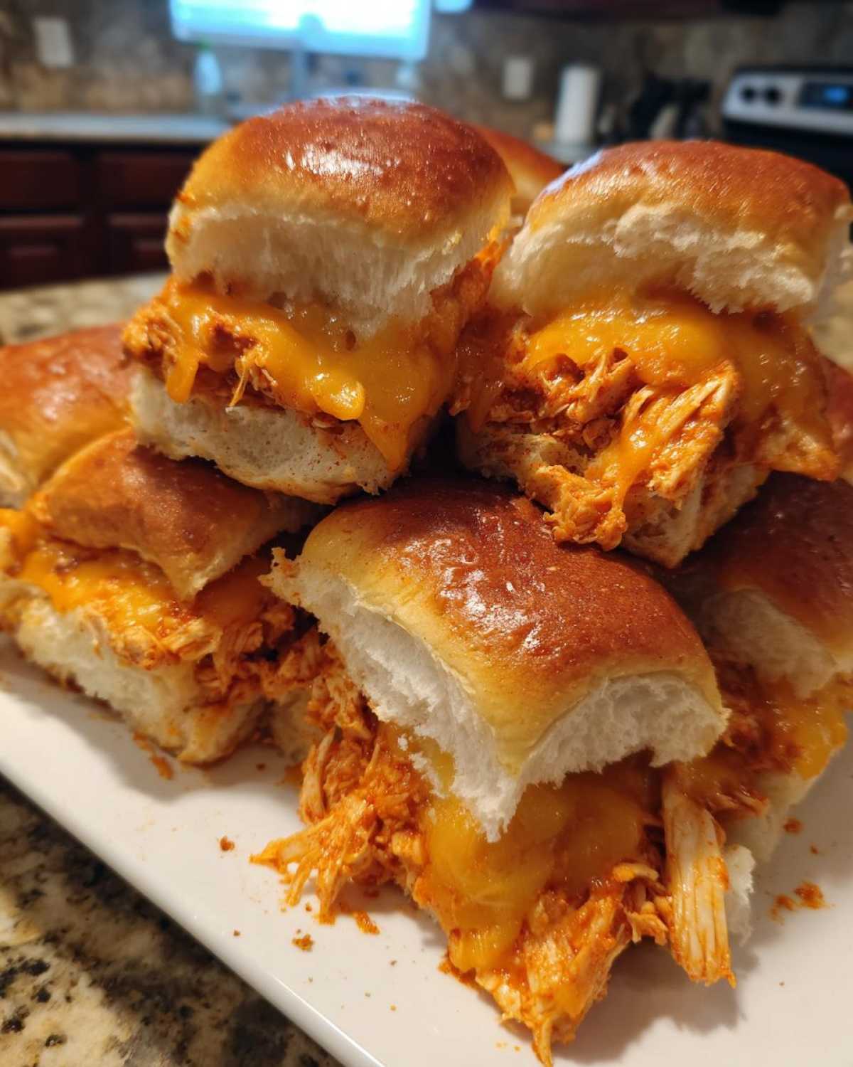 A close-up stack of freshly baked buffalo chicken sliders featuring shredded chicken, melted cheddar cheese, and shiny rolls.