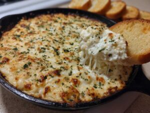 A scoop of cheesy garlic bread dip being lifted from a cast-iron skillet, showing a dramatic cheese pull, served with toasted bread slices.